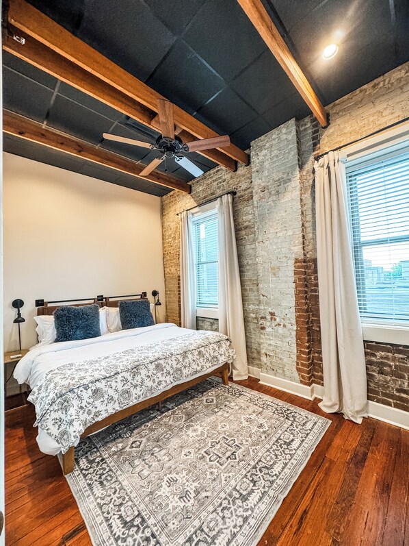 A bedroom with a bed, a ceiling fan, a window with curtains, and a brick wall.