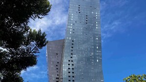 A tall, modern building with a reflective glass facade, surrounded by trees and a smaller adjacent building.