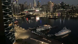 A waterfront cityscape with modern buildings, a marina with yachts, and a pedestrian walkway along the water.