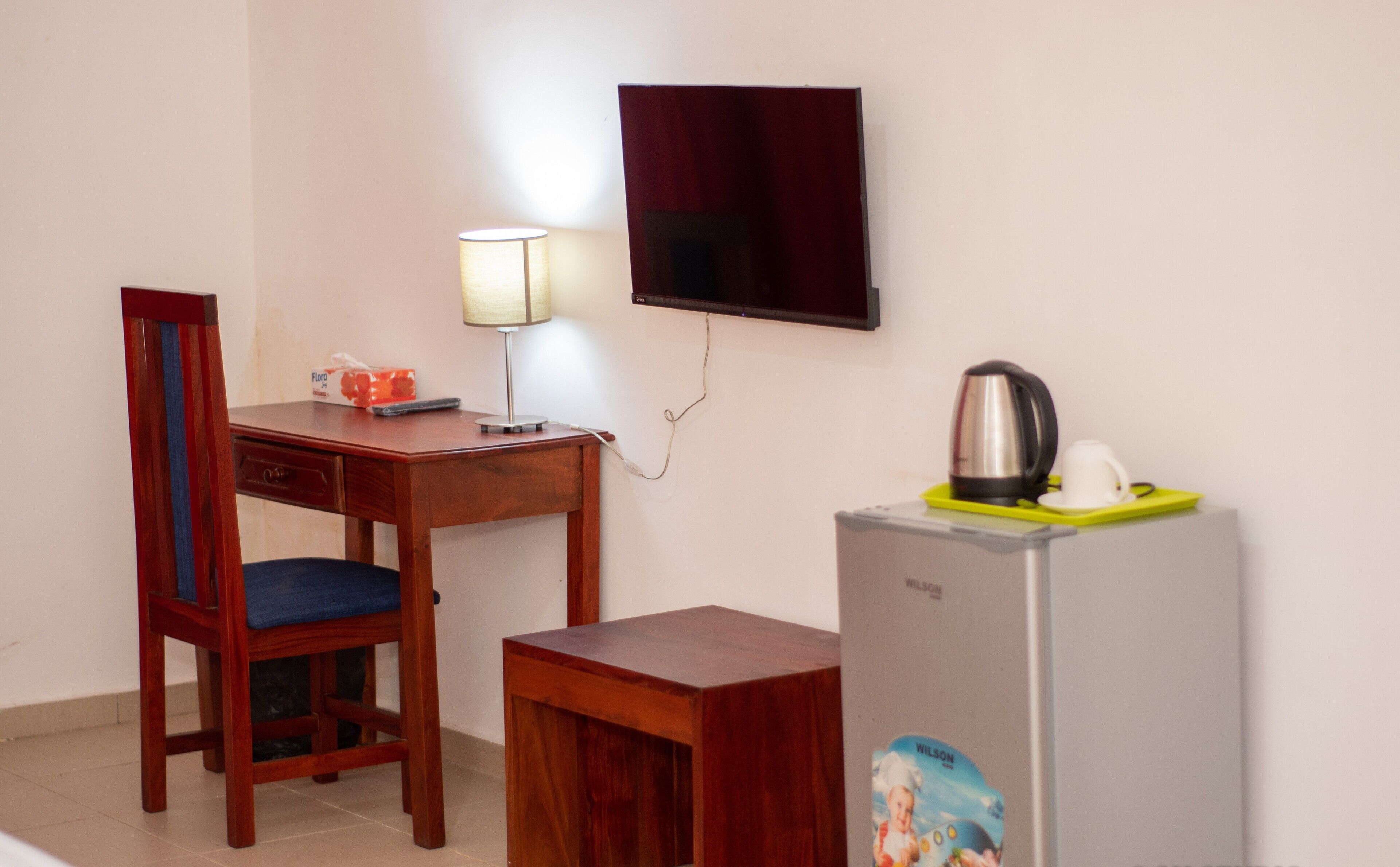 A small room with a wooden desk, a chair, a television mounted on the wall, and a refrigerator with a kettle and cup on top.