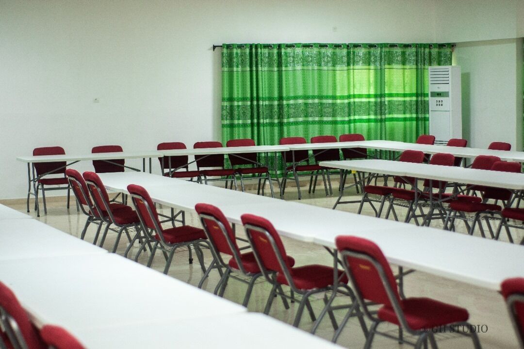 A conference room with long tables and red chairs, a green curtain, and a white air conditioning unit.