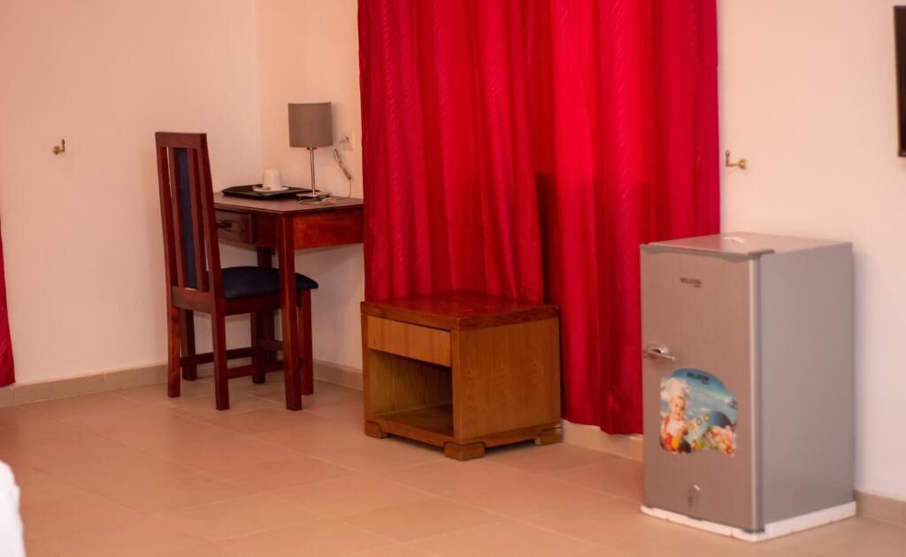 A small room with a wooden chair, a desk with a lamp, a red curtain, and a refrigerator.