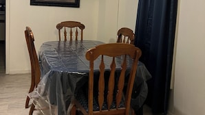 A dining area with a table covered by a plastic sheet, wooden chairs, a ceiling fan, a framed picture on the wall, and a dark curtain.