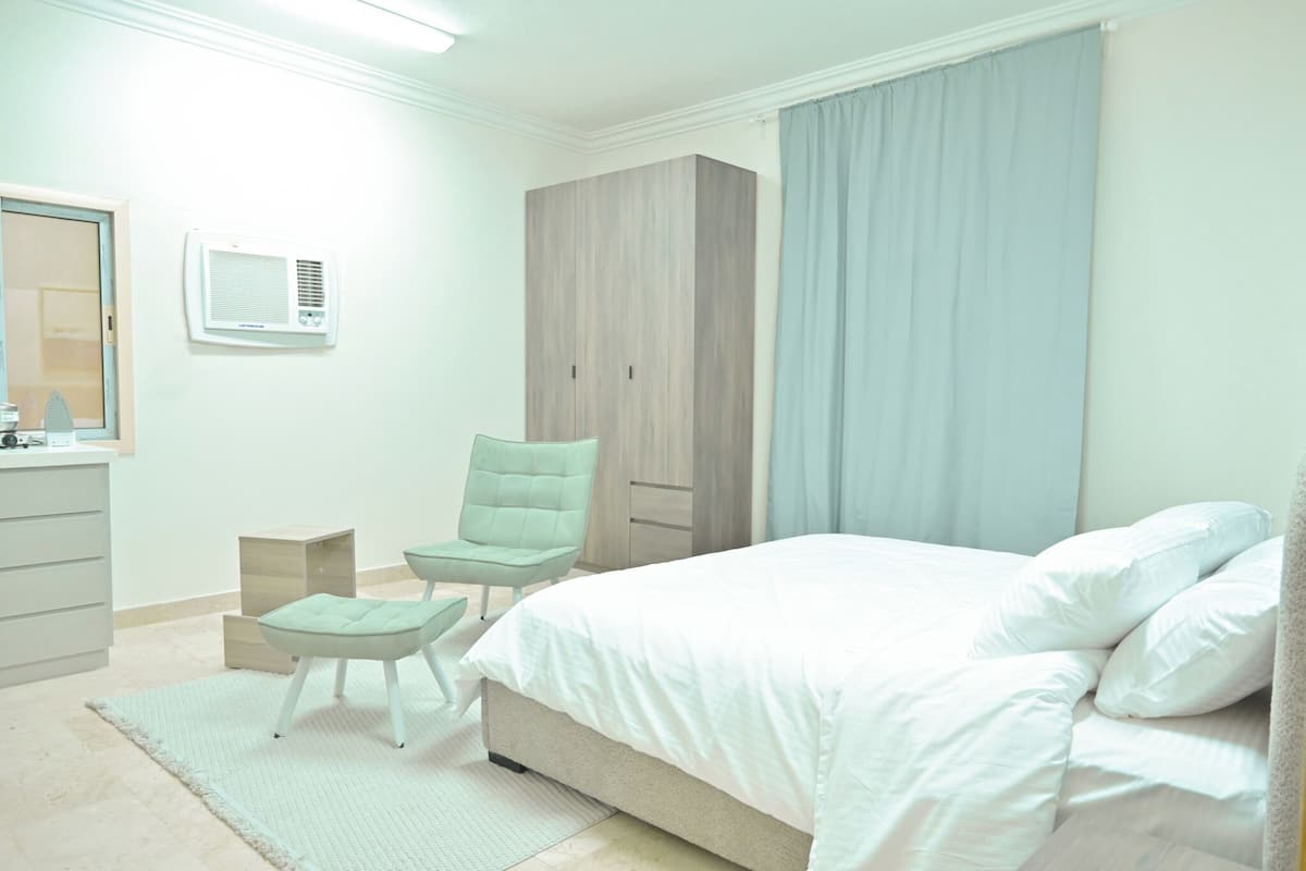 A bedroom with a bed, a chair, a wardrobe, a mirror, and an air conditioning unit.