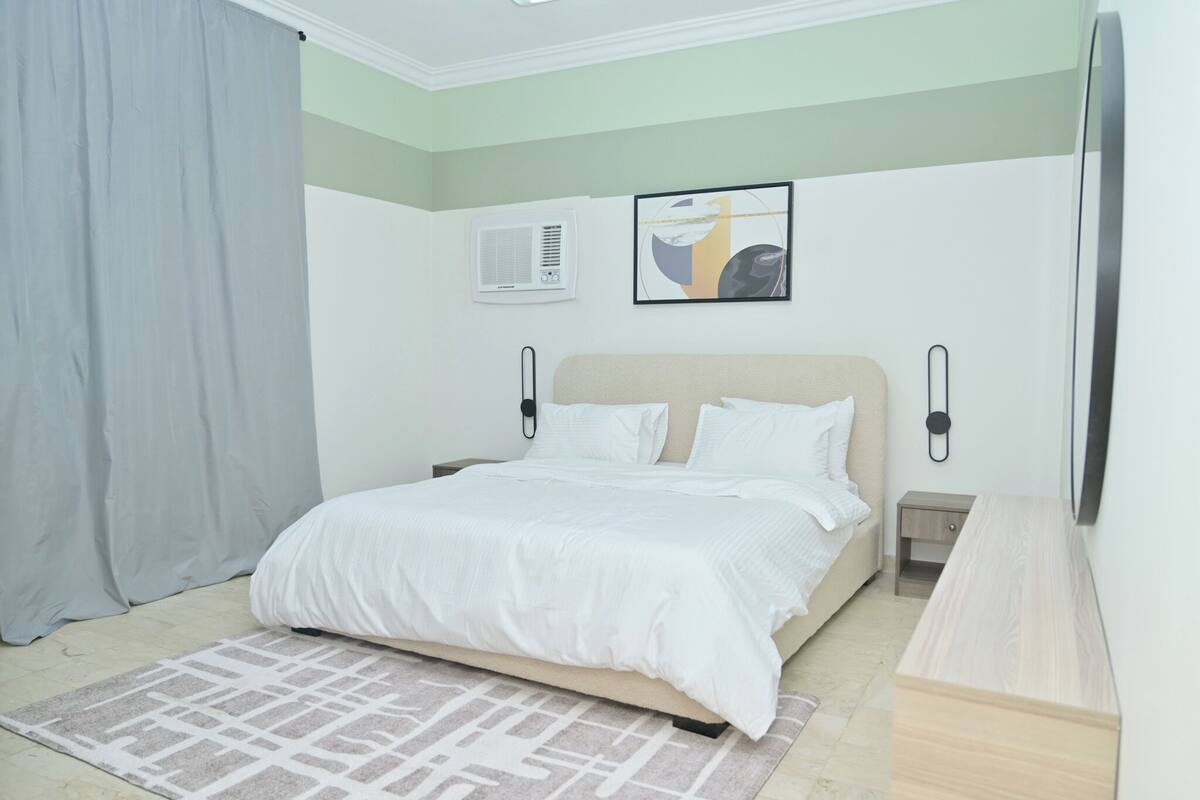 A bedroom with a bed, a bedside table, a nightstand, a wall-mounted air conditioner, and a framed picture on the wall.