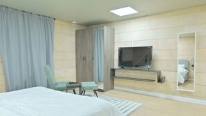 Moderni hotellihuone, jossa on sänky, seinälle asennettu televisio, peili sekä oleskelualue, jossa on tuoli ja pöytä.