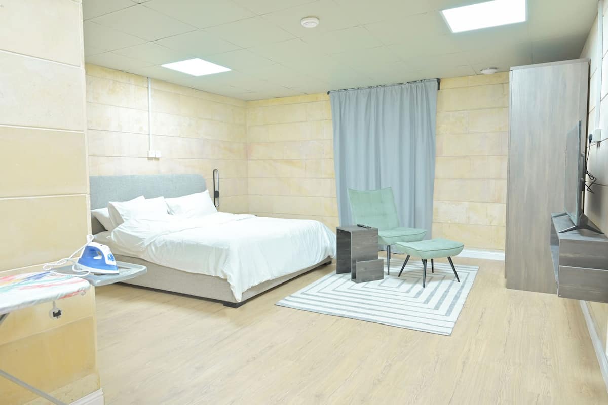 A modern hotel room with a bed, a small table with an ironing board, a chair, a TV, and a bathroom area.