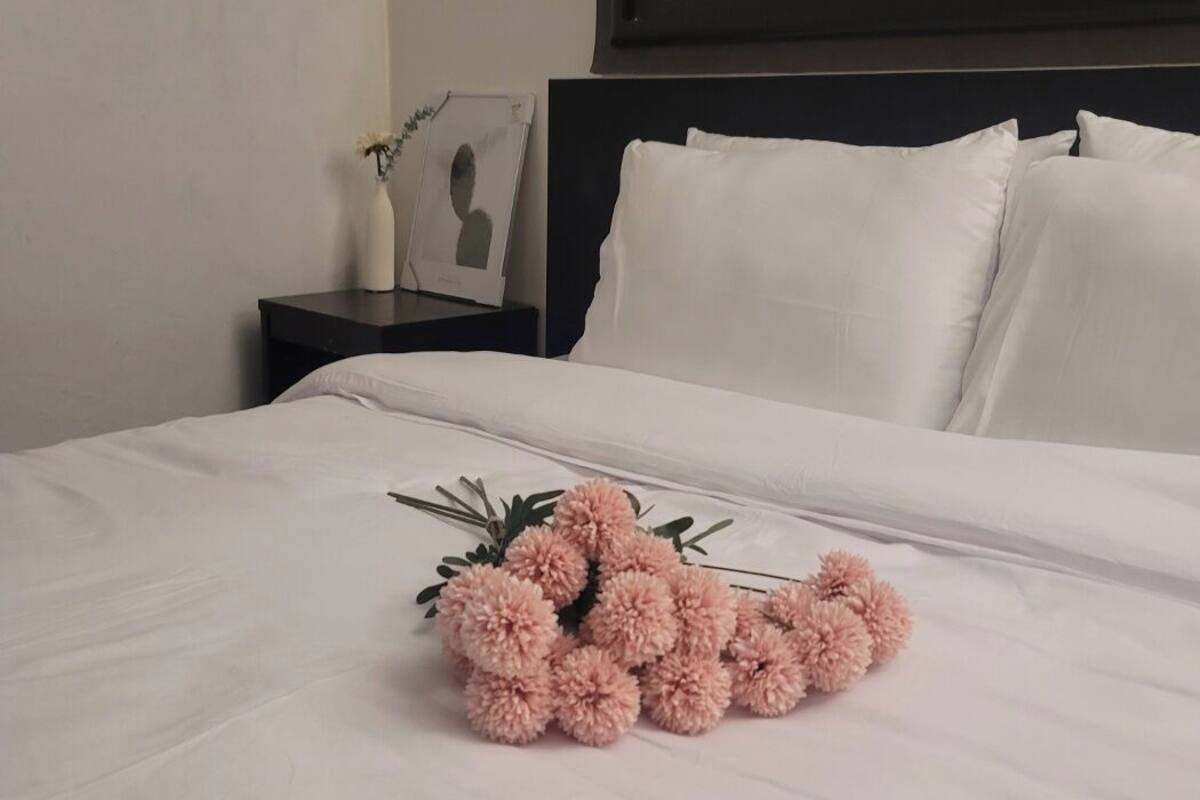 A neatly made bed with white linens, a dark headboard, and a small bedside table with a vase and a framed photo.