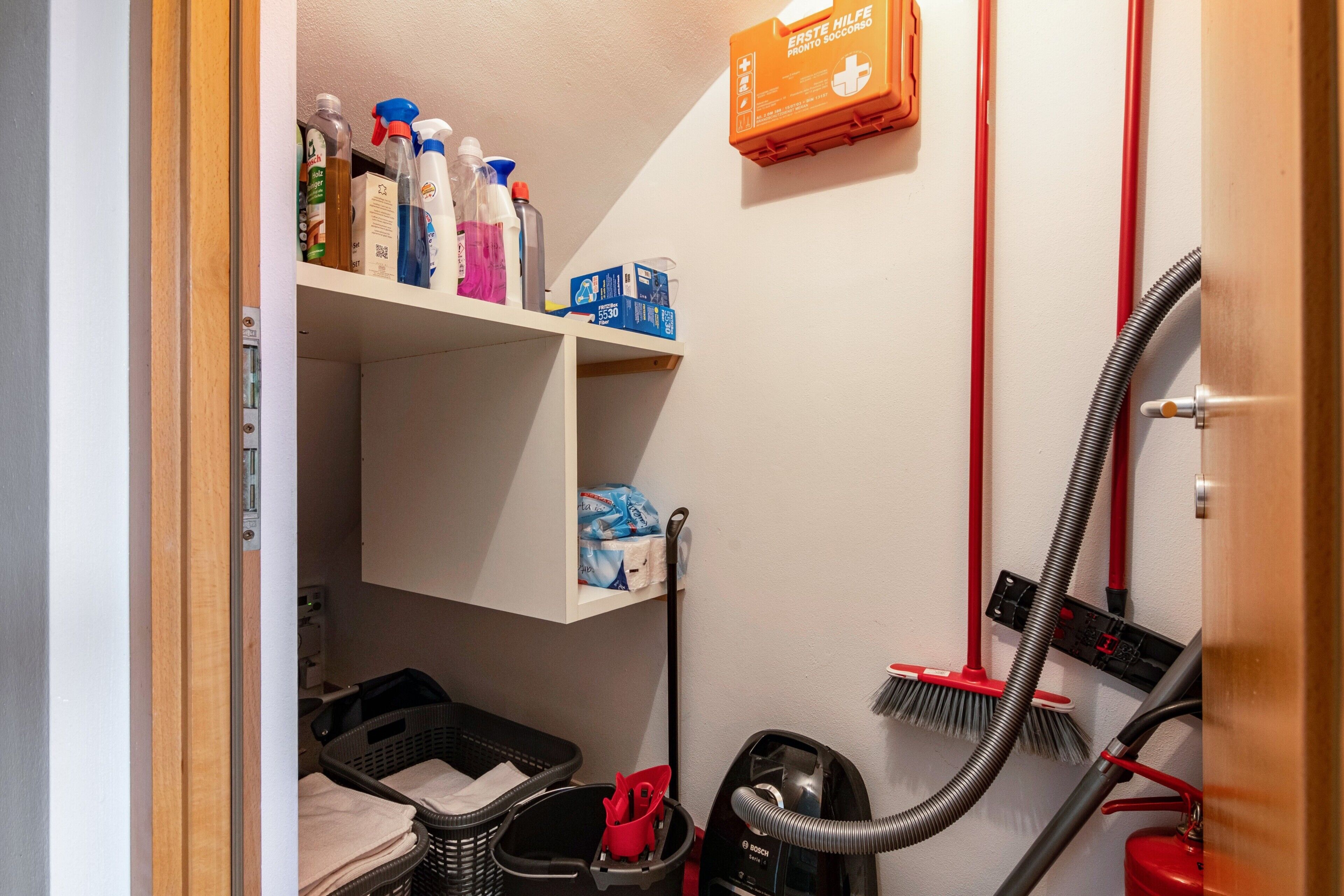 Een wasserij met een Bosch stofzuiger, een rode stofzuigerslang, een bezem en diverse schoonmaakmiddelen op planken.