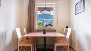 Un comedor con una mesa, sillas, una planta en maceta, un ventanal con vistas a montañas y una lámpara colgante.