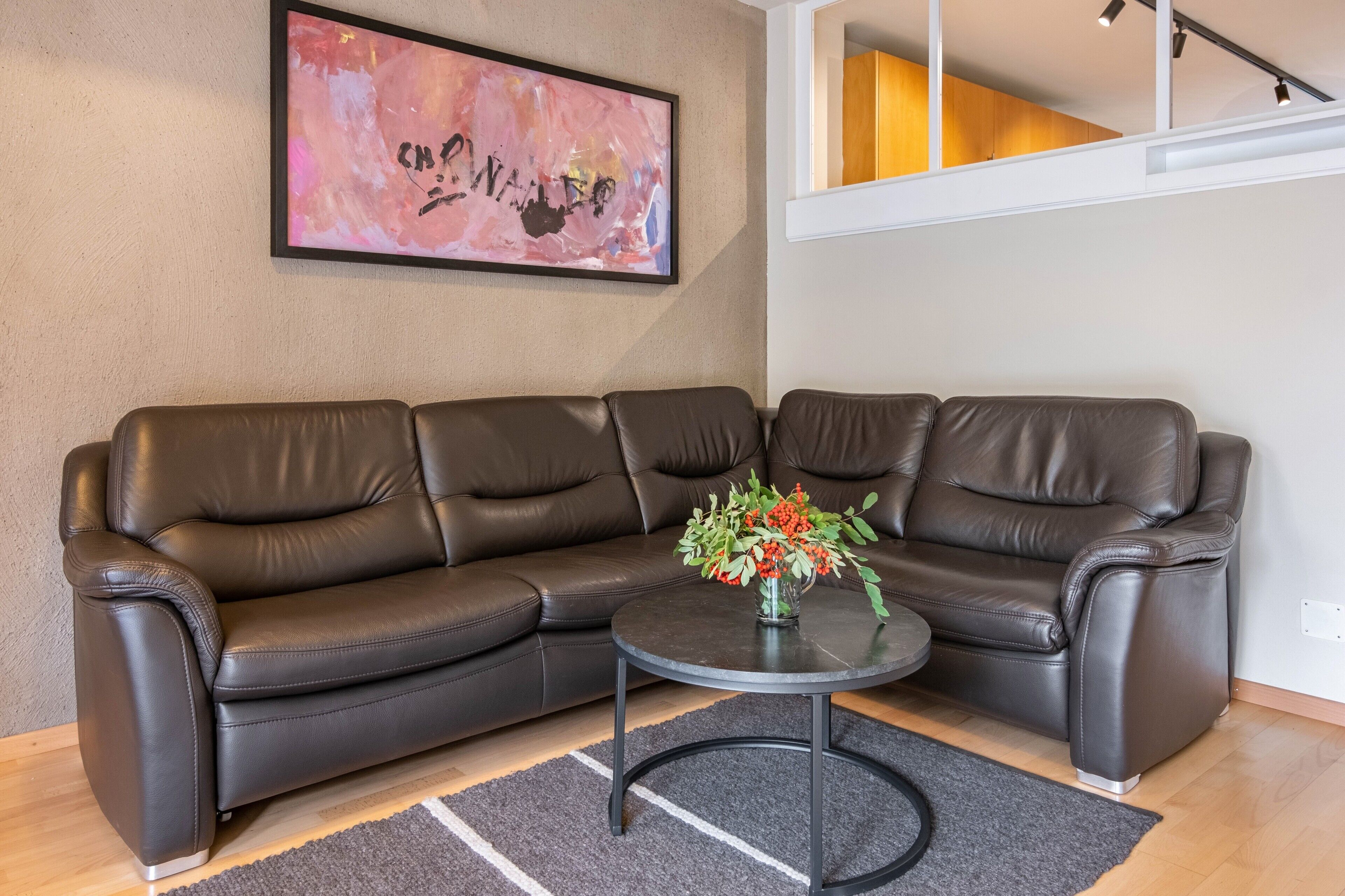 A brown leather sofa, a small round table with a vase of flowers, and a framed abstract painting on the wall.