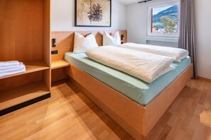 A hotel room with a wooden bed, a bedside table, a shelf with towels, a wall-mounted artwork, and a window with a view of a mountain and buildings.