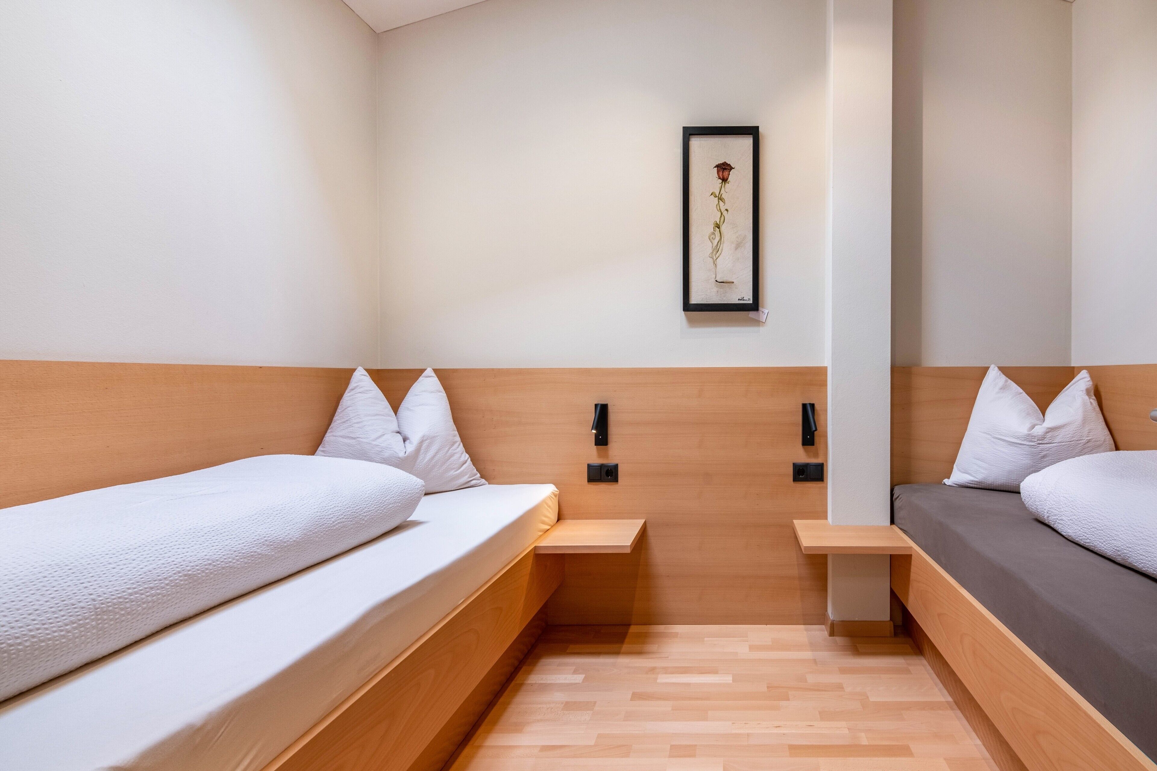 A modern hotel room with two beds, a wooden headboard, a small bedside table, a framed artwork on the wall, and a light-colored floor.