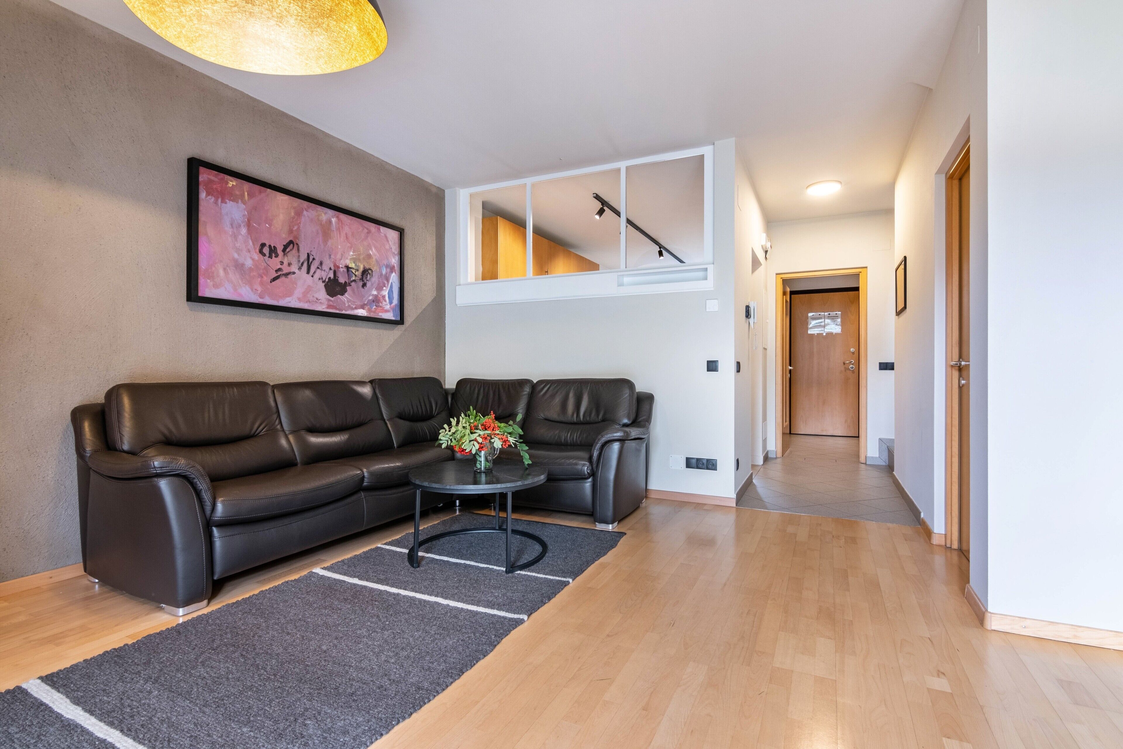 A modern living room with a leather sofa, a coffee table with flowers, a staircase, and a hallway leading to a door.