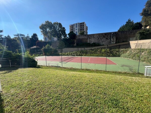 A tennis court with a green and red surface, surrounded by a chain-link fence, with a stone wall and trees in the background.