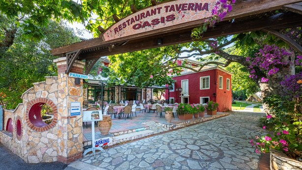 Ein steinernes Eingangstor mit einem Schild für Restaurant Tavern Oasis, Außenbereich zum Sitzen und ein rotes Gebäude im Hintergrund.