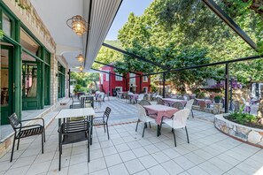 Un patio al aire libre con mesas y sillas, un edificio rojo al fondo y un árbol grande que da sombra.