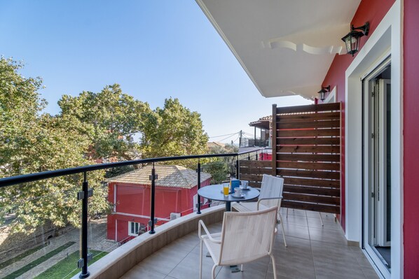 Un balcón con una mesa y sillas, un edificio rojo al fondo y vistas a árboles.