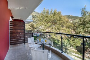 Un balcón con sillas de mimbre blanco y una pequeña mesa redonda, con vistas a un paisaje verde con árboles y una colina.