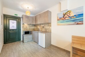 Eine kompakte Küche mit einer Mikrowelle, einem kleinen Kühlschrank und einem Spülbecken. Eine grüne Tür mit dekorativem Glasfenster und einem Ausgangsschild. An der Wand hängt ein Gemälde von einer Küstenlandschaft.