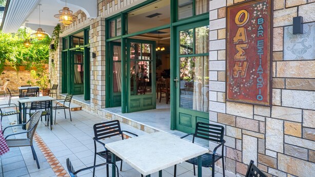 Ein Straßencafé mit Tischen und Stühlen, eine Steinmauer und ein Schild mit der Aufschrift 'OASTRO BAR ESTIA TOPIO'.