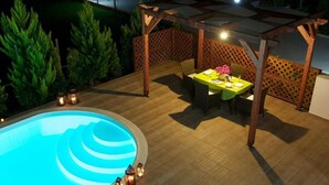 Una piscina con muelle de madera, zona de comedor cubierta y estructura de pérgola.