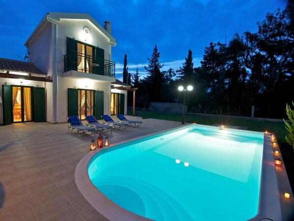 Una casa de dos pisos con piscina, mobiliario exterior y un patio bien iluminado.
