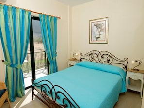A bedroom with a bed, a nightstand, a framed picture on the wall, and a balcony with curtains.