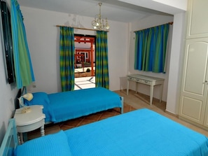 A bedroom with two beds, a desk, a chair, a lamp, and a view of the outside.