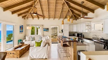 A spacious living area with a wooden ceiling, a sofa set, a kitchen with white cabinets, and a view of the ocean through large windows.