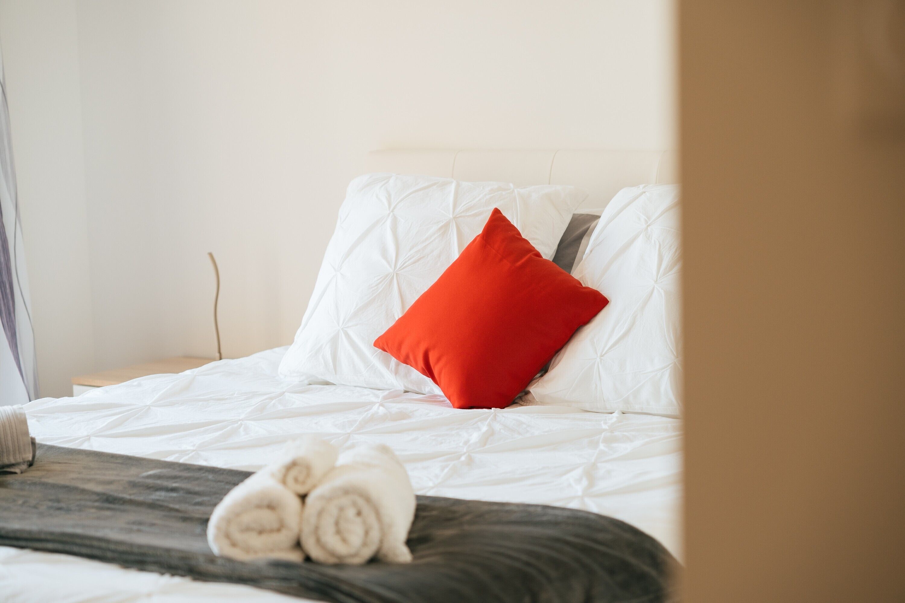 A neatly made bed with white bedding and a red pillow, a wooden headboard, and a rolled towel on the bed.