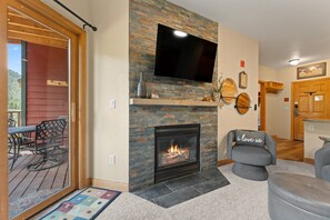A living room with a stone fireplace, a flat-screen TV mounted above it, and a cozy seating area with a grey sofa and ottoman. - Enjoy Stunning Mountain Views from the Private Balcony & Convenient Location! (Keystone)