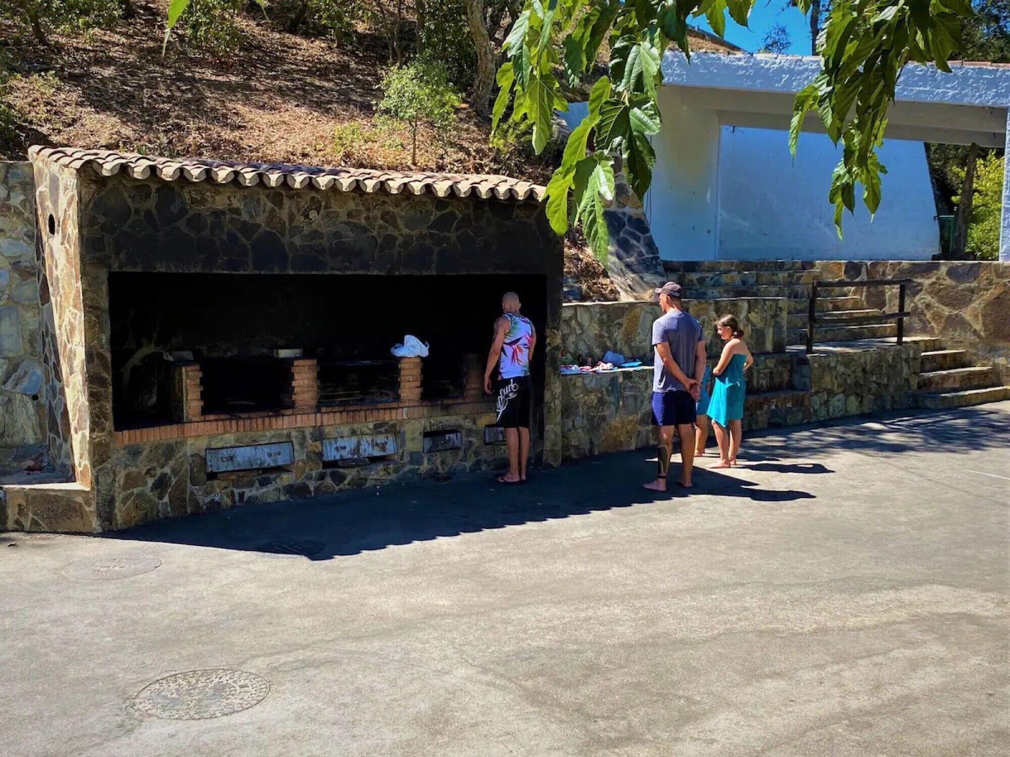 Zona de barbacoa de piedra con techo de tejas, rodeada por un muro de piedra y escalones que conducen a un edificio.