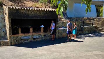 Zona de barbacoa de piedra con techo de tejas, rodeada por un muro de piedra y escalones que conducen a un edificio.