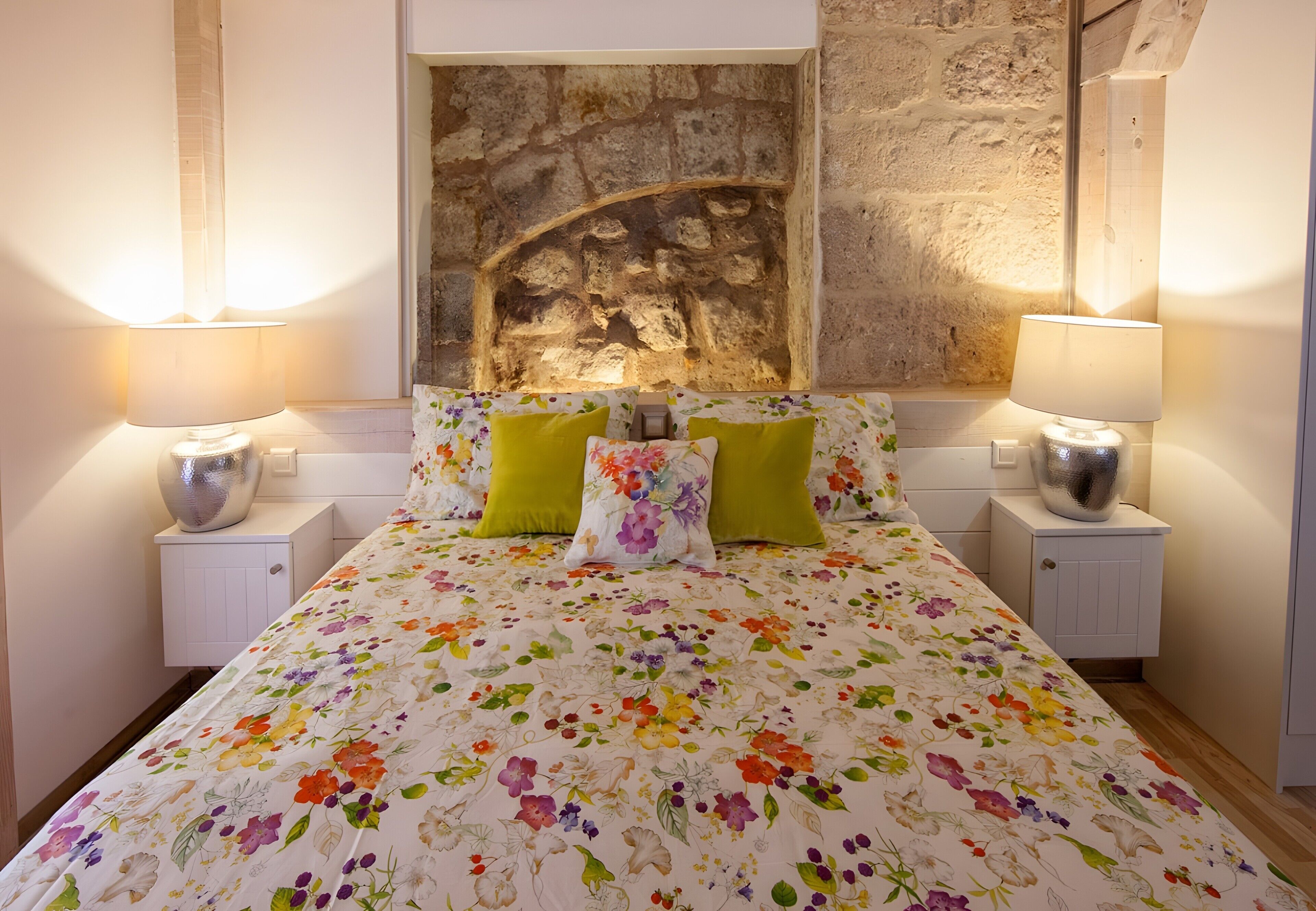 Ein Schlafzimmer mit floralem Bettbezug, zwei Nachttischen mit Lampen, einer Steinwand und einem Bett mit zwei Kissen.