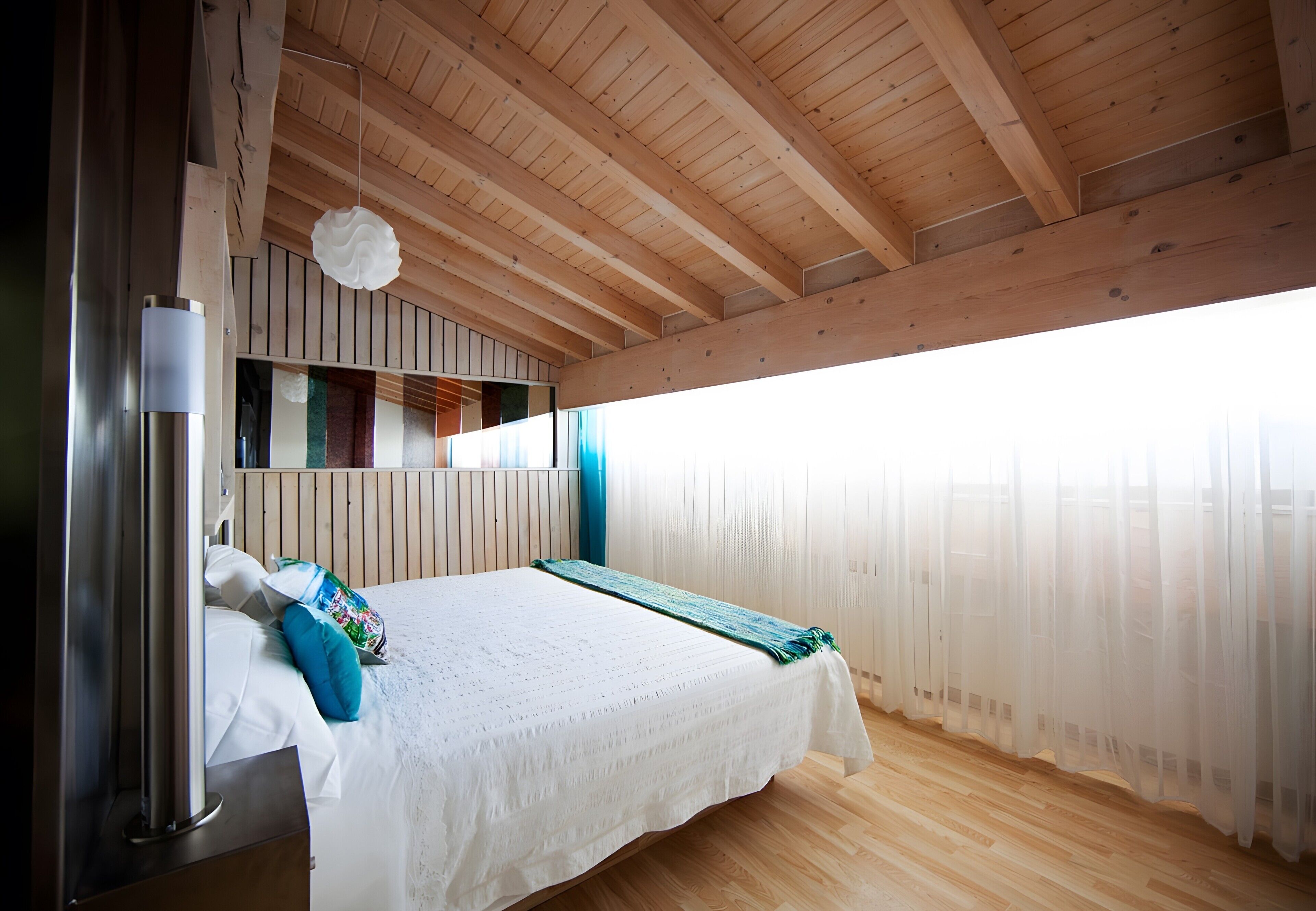 A bedroom with a wooden ceiling, a bed with white bedding, a bedside table, and a window with sheer curtains.