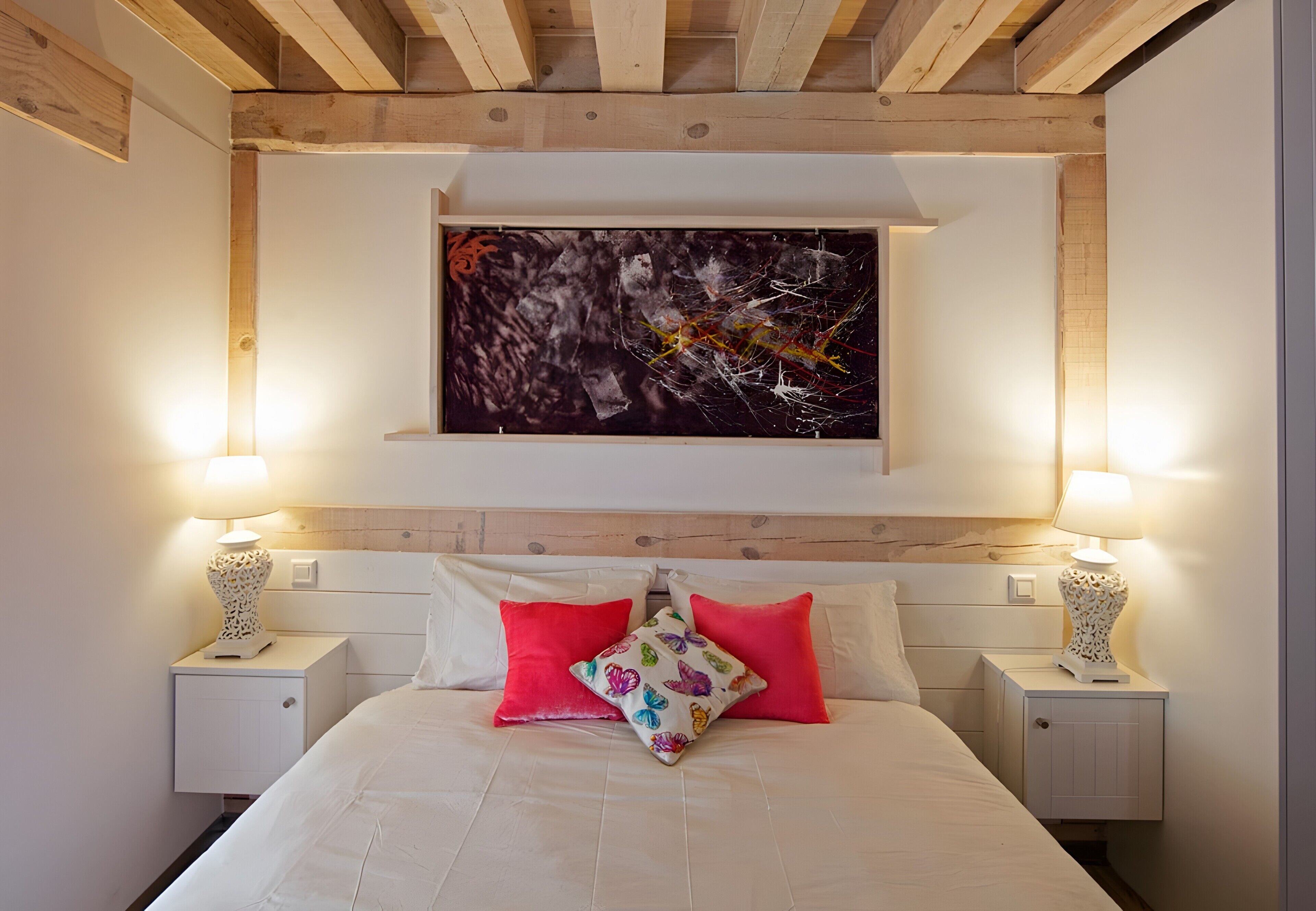 Un dormitorio con cama, dos mesitas de noche, una obra de arte colgada en la pared y techo de madera.
