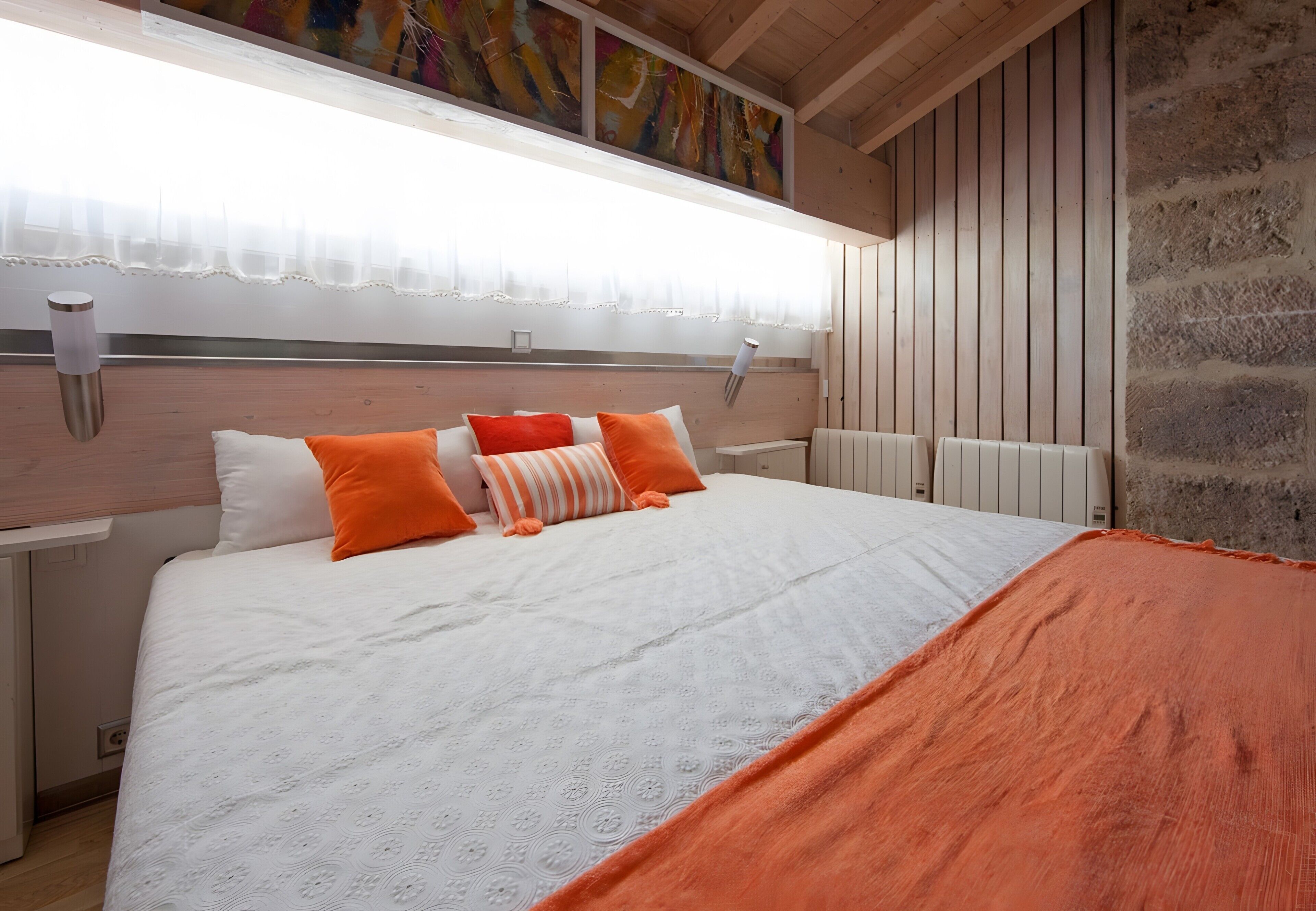 A bedroom with a large bed, white bedding, orange pillows, a white headboard, a wooden ceiling, and a stone wall.