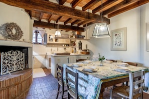 Een eetruimte met een tafel voor vier personen, houten stoelen, een open haard en een keuken op de achtergrond.