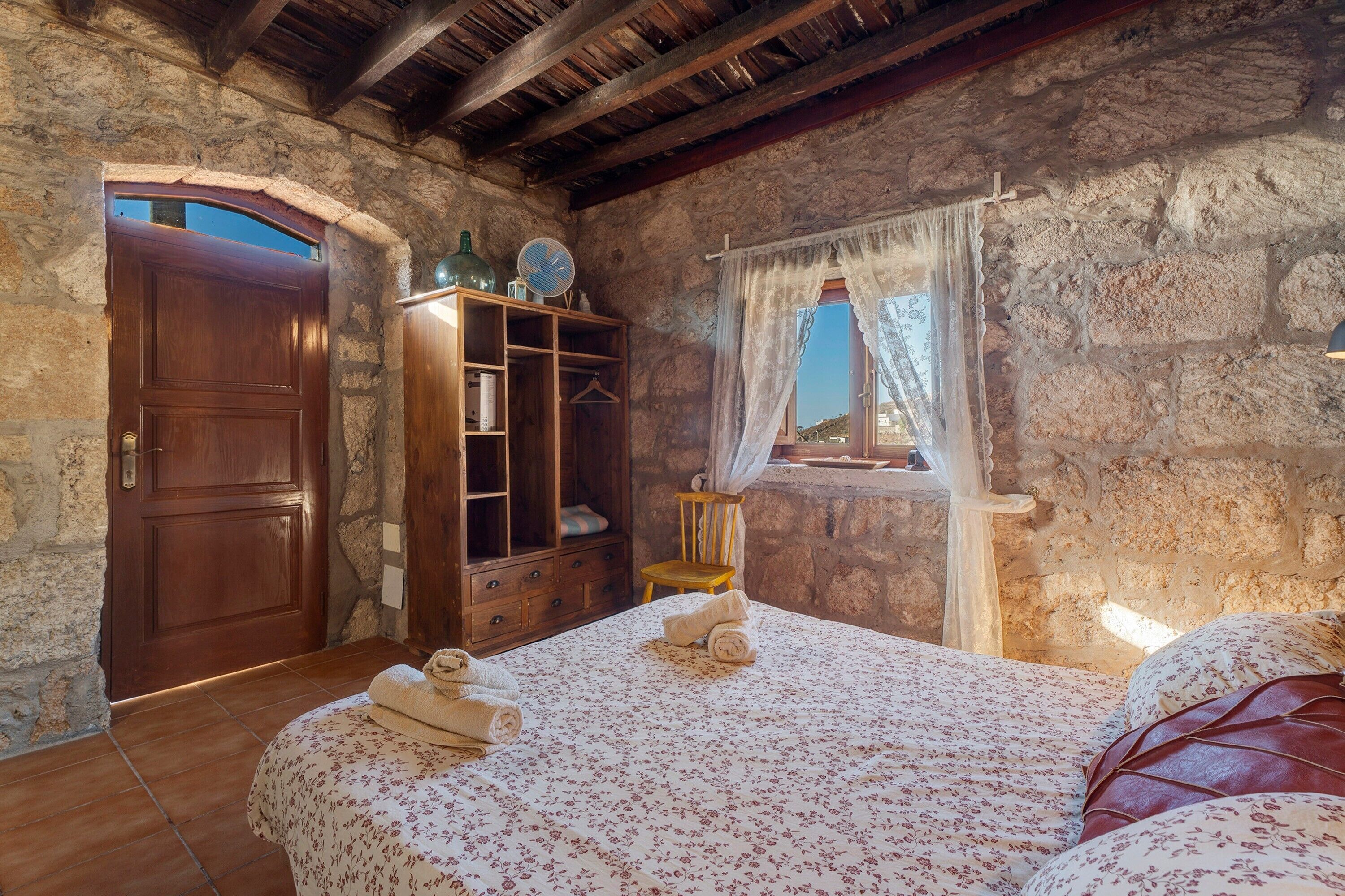 A rustic room with a wooden door, a wooden cabinet, a bed with floral bedding, and a window with a view.