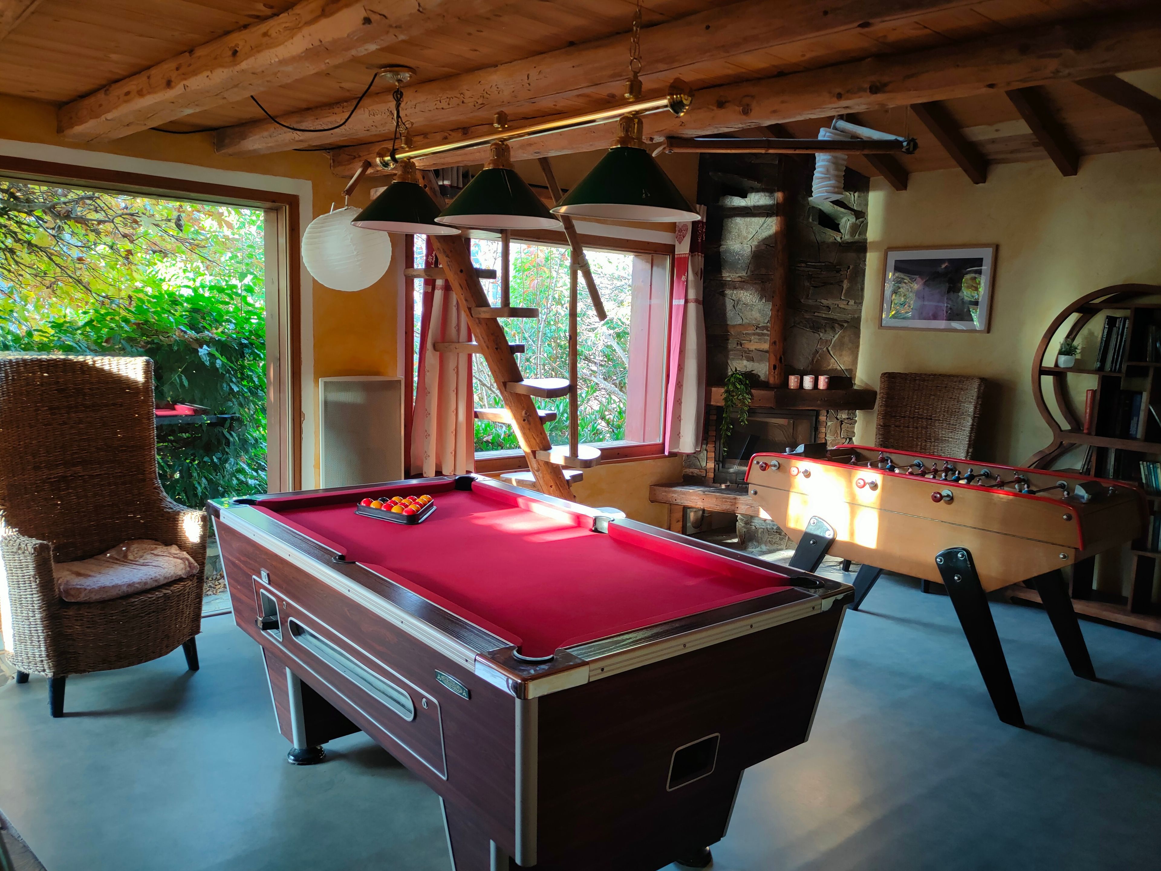 A pool table with a red felt surface, a foosball table, a wicker chair, and a bookshelf with various items.