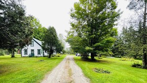 Un chemin de campagne mène à une maison blanche avec une porte verte, entourée d’une végétation luxuriante et d’arbres.