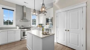 A modern kitchen with white cabinetry, a central island, stainless steel appliances, pendant lighting, and a view of the outdoors. - Charming 3 bedroom beach house in Cape Charles, VA  (Cape charles)