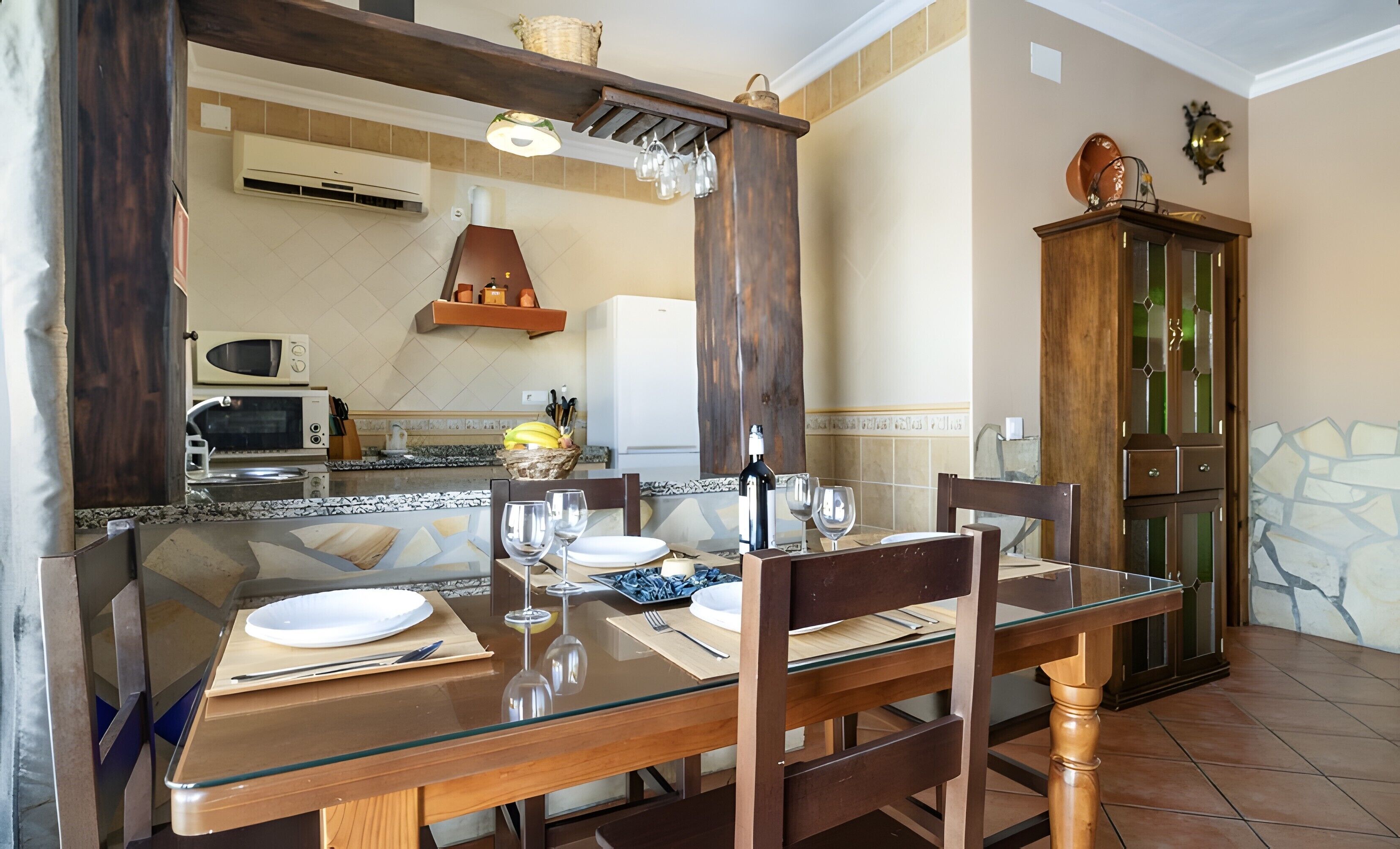 A dining area with a glass table, wooden chairs, a wooden cabinet, and a kitchen in the background.