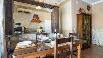 A dining area with a glass table, wooden chairs, a wooden cabinet, and a kitchen in the background.