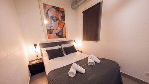 A neatly made bed with a dark bedspread and two pillows, a nightstand with a lamp, and a painting above the bed.