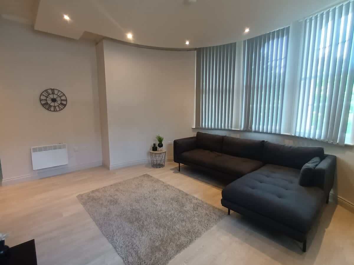 A modern living room with a sectional sofa, a round clock, a small side table with a plant, and a large window with blinds.