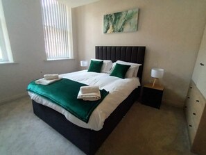 A neatly made bed with a green throw blanket, pillows, and two folded towels. There is a bedside table with a lamp, a painting on the wall, and a window with blinds. - Elegant 1Bed Apt in Leeds City Centre (Leeds)
