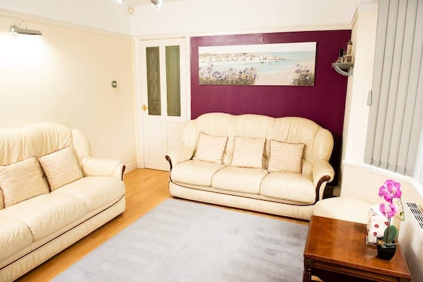 Una sala de estar con dos sofás color crema, una alfombra gris, una mesa de centro de madera y una pared morada con una pintura costera.