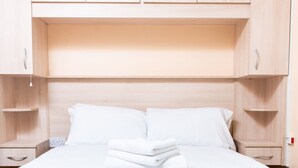 Una cama bien hecha con sábanas blancas y una pila de toallas dobladas, un cabecero de madera y armarios de madera de color claro sobre la cama.